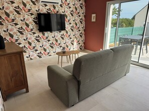 A living room with a grey sofa, a wooden cabinet, a small round table, and a TV mounted on the wall. - Les Parures d’Anthésim 20 : Cotentin – A/C & Green'Up EV Charger (Saint-Floxel)