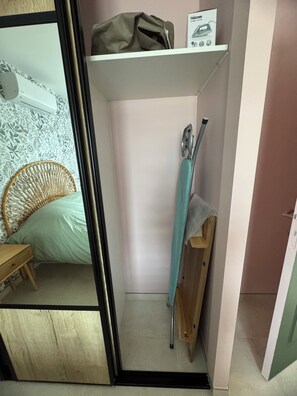 Una habitación pequeña y funcional con cama, una silla, un estante con una bolsa y una caja, y una puerta.