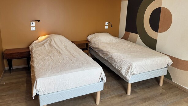 A room with two single beds, a wooden nightstand, a wall-mounted air conditioner, and a geometric wall design. - Les Parures d’Anthésim 20 : Cotentin – A/C & Green'Up EV Charger (Saint-Floxel)