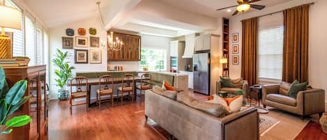 A spacious living area with a large sofa, a dining table with chairs, a kitchen area with a refrigerator, and a ceiling fan.