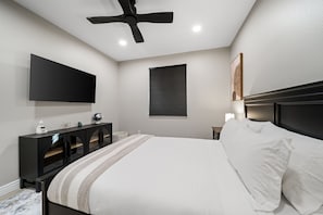 Ein Schlafzimmer mit einem Bett, einem an der Wand montierten Fernseher, einem Deckenventilator, einem Fenster mit Jalousien und einem Nachttisch mit Lampe.
