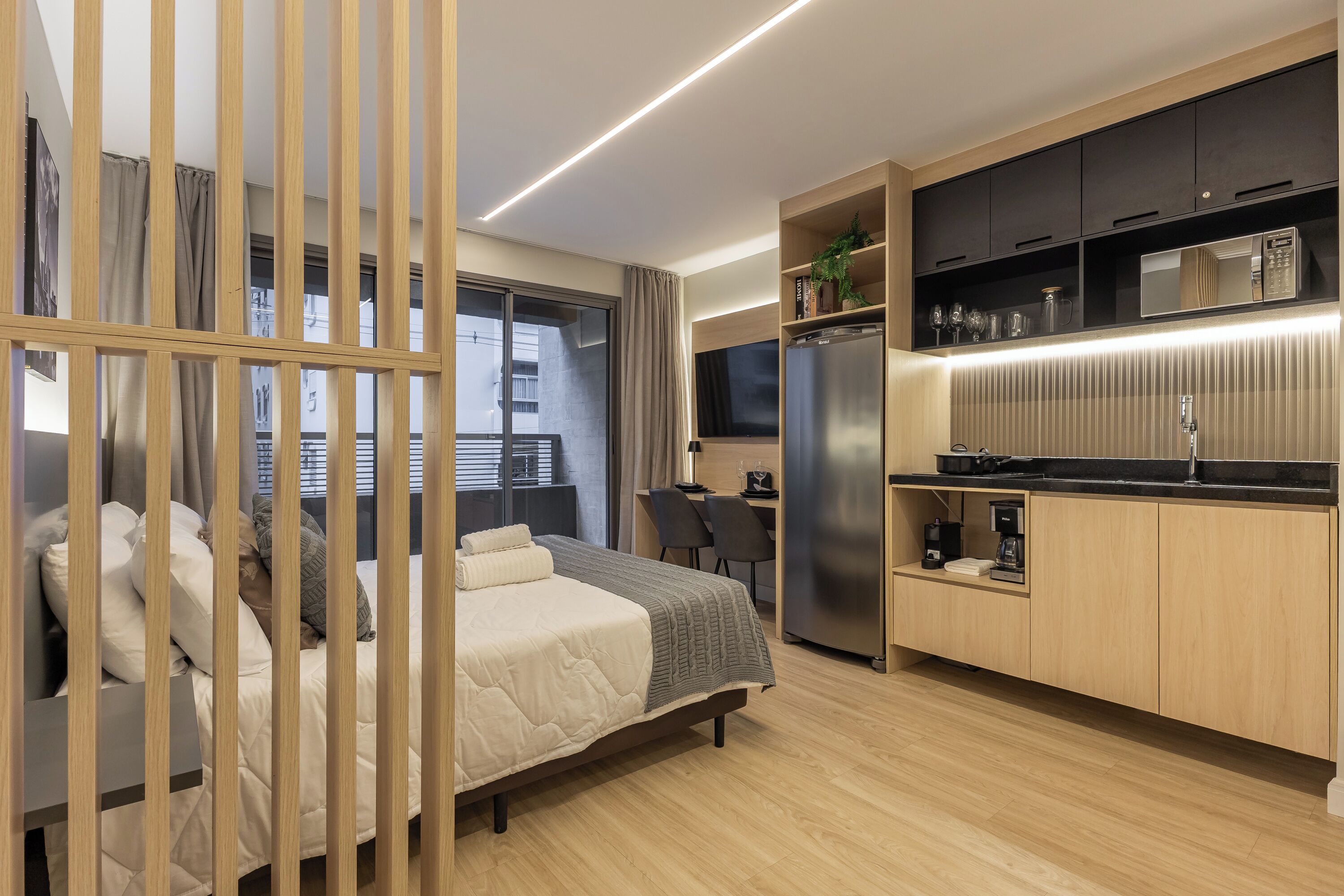 A modern hotel room with a bed, a desk, a kitchenette, and a large window.