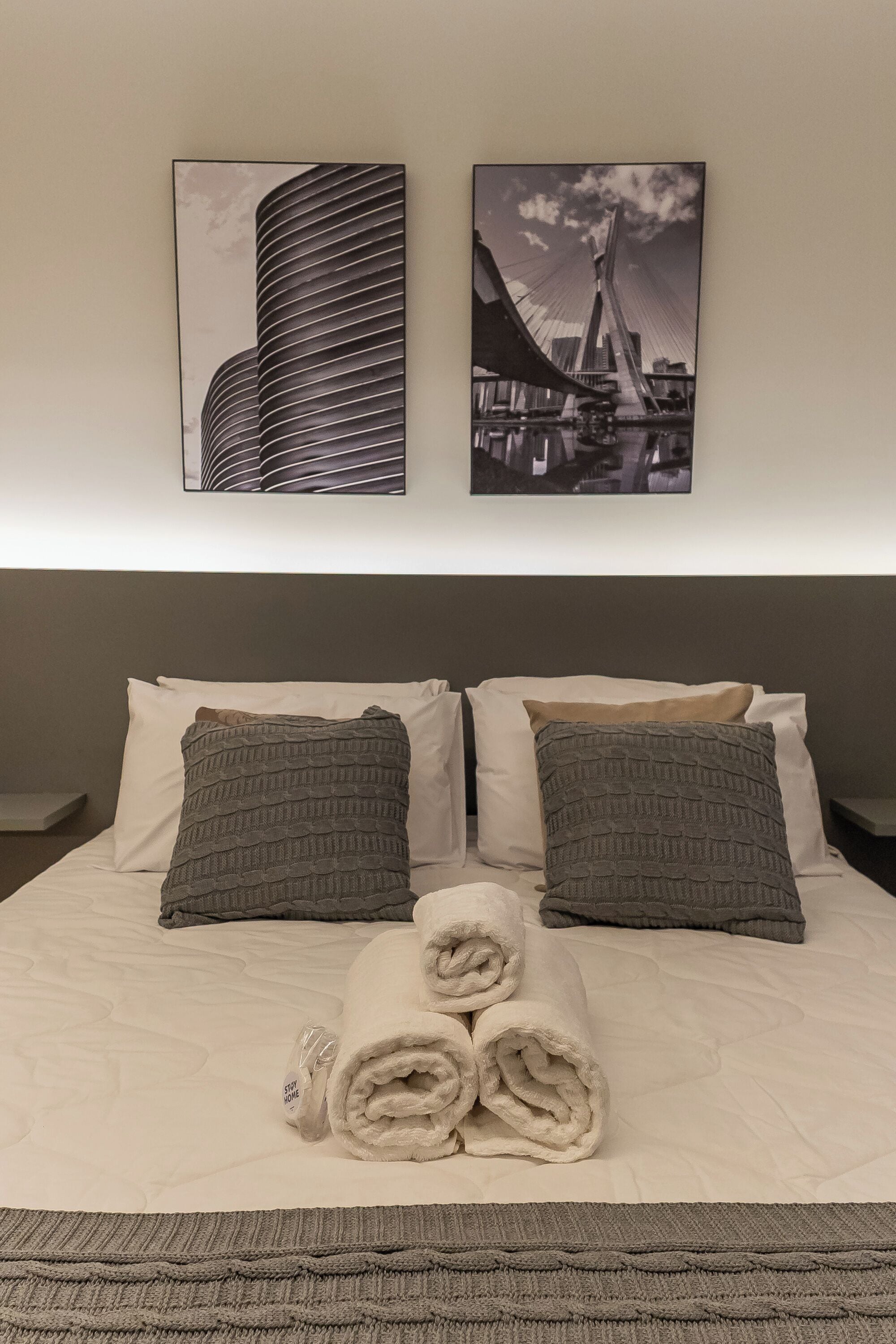 Habitación de hotel con una cama grande, dos almohadas, una toalla enrollada y dos fotografías en blanco y negro de un paisaje urbano en la pared.