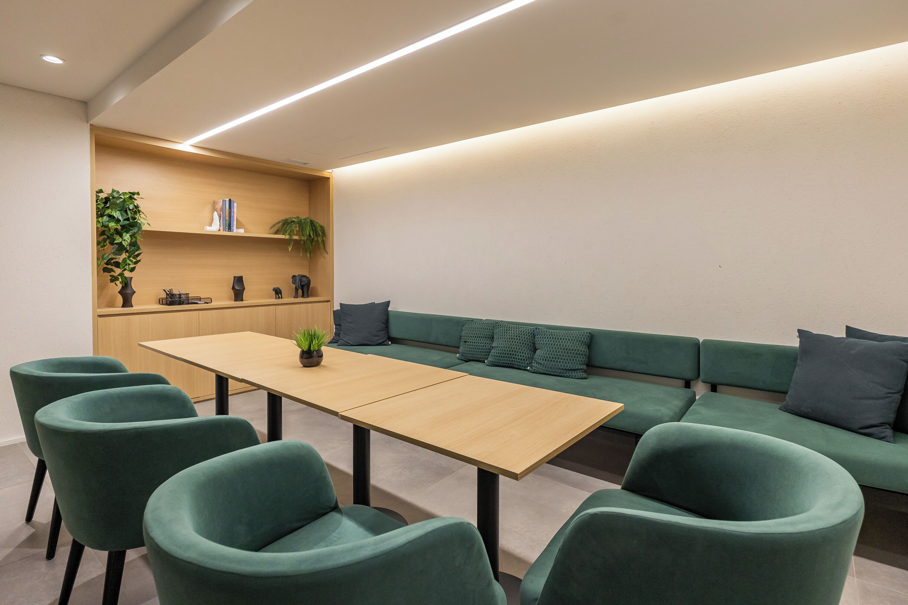 Una sala de reuniones moderna con una larga mesa, sillas tapizadas en verde, un sofá con cojines y una estantería de madera con plantas y libros.