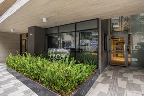 Entrada de un edificio moderno con puerta de vidrio, un cartel que dice 'Oscar' y una jardinera con plantas verdes.