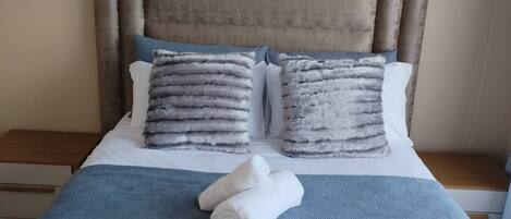 A neatly made bed with a headboard, two pillows, and rolled towels on a blue bedspread.