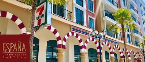 Un edificio de varios pisos con una destacada tienda de conveniencia 7-Eleven en la planta baja, un letrero que dice 'ESPANA CONDO RESORT PATTAYA' y una serie de arcos con diseño a rayas.