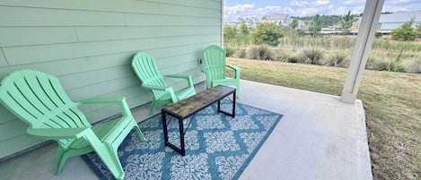 A covered porch with three green Adirondack chairs and a small wooden table, a blue patterned rug, and a view of a residential area with houses and greenery.