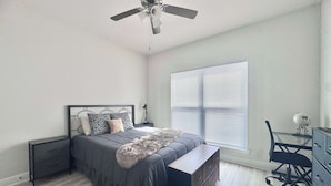 A bedroom with a bed, bedside tables, a desk, a chair, a ceiling fan, and a window with blinds. - Coastal Escape in Sea Grove (Port St. Joe)