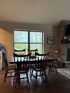 A dining area with a table and chairs, a stone fireplace, and a large window with a view of trees.