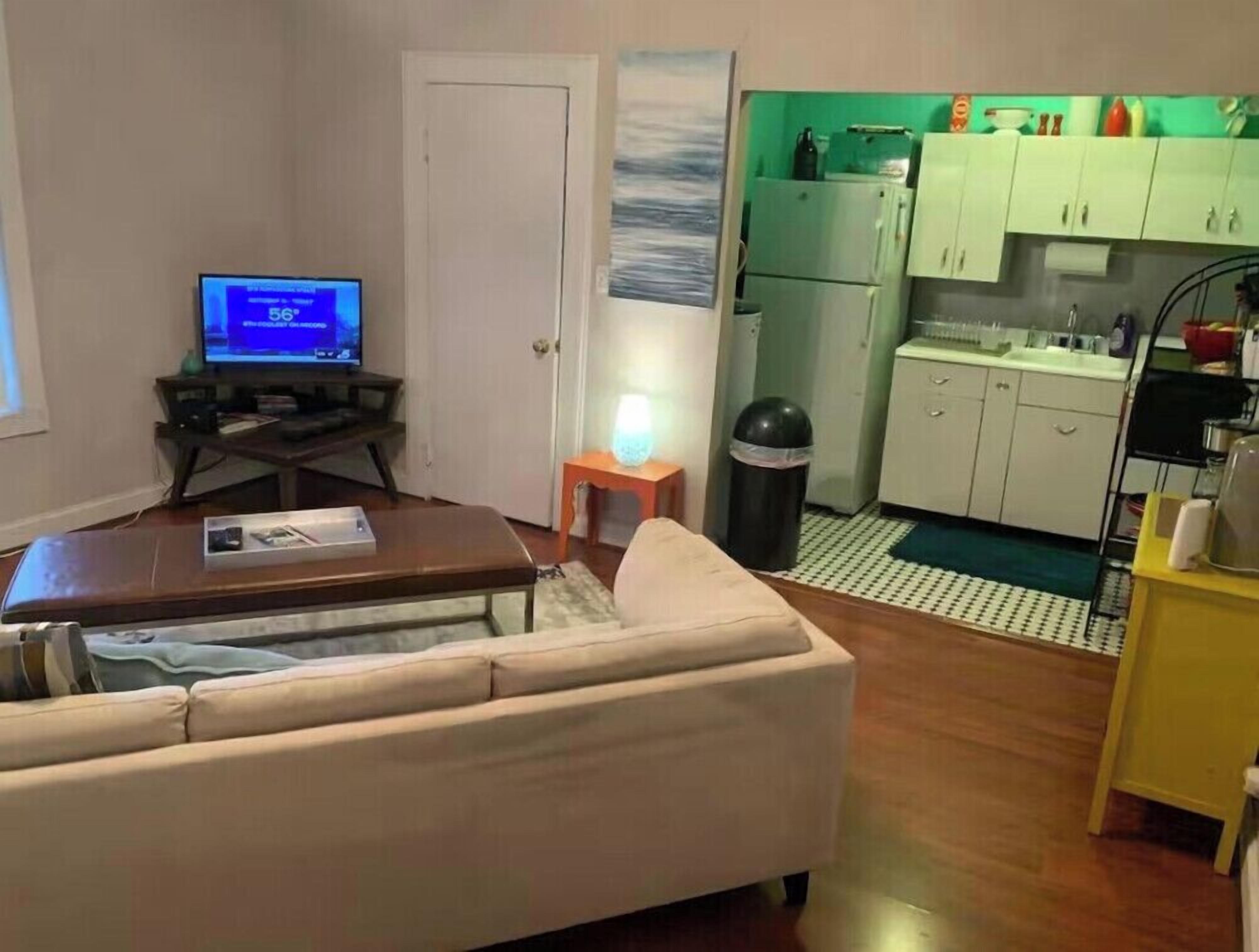 A living room with a sofa, a television, a small table, and a kitchen area with cabinets and a refrigerator.