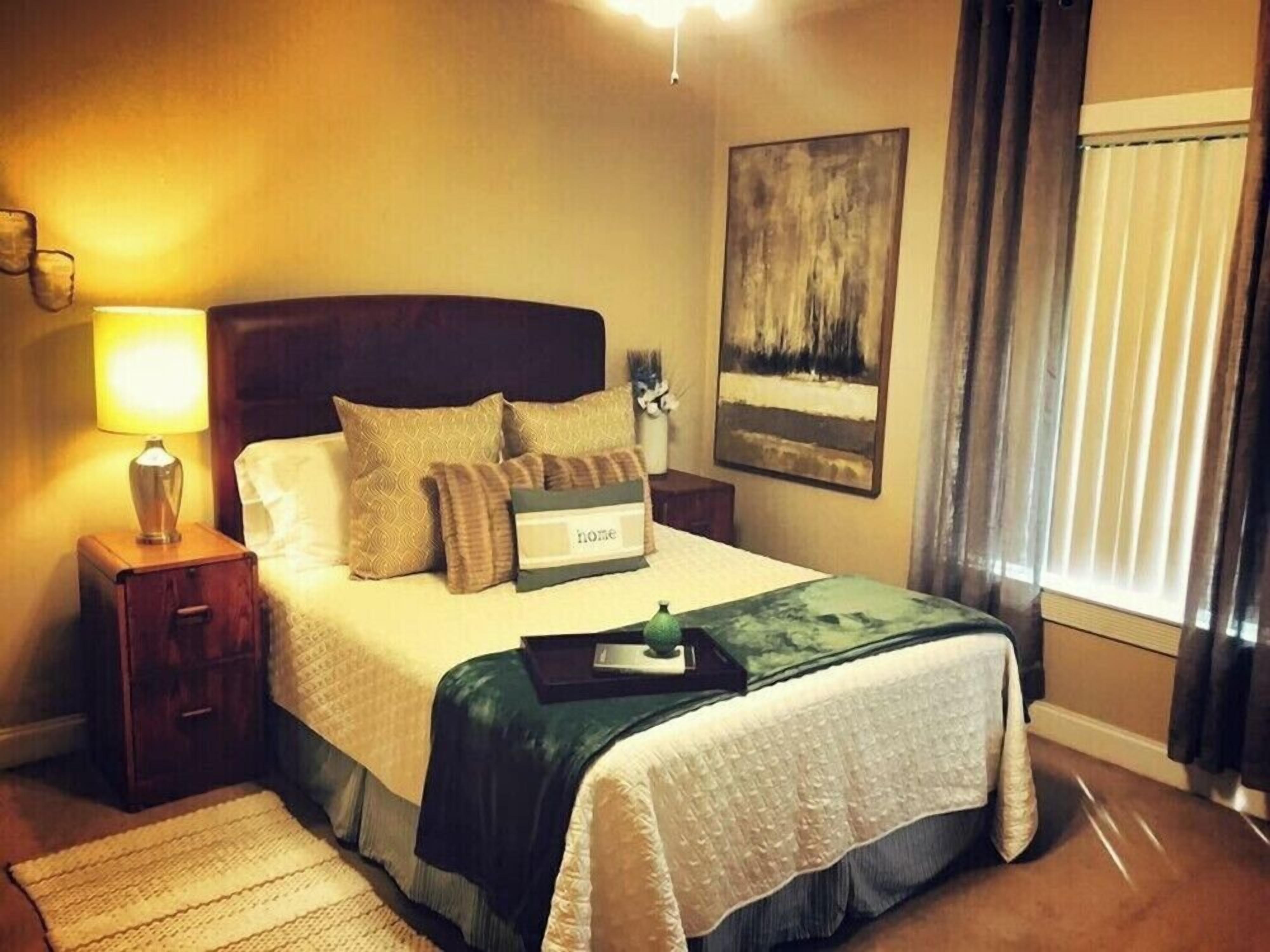 Een hotelkamer met een bed, een nachtkastje met een lamp, een lijst aan de muur en een raam met gordijnen.