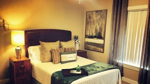 Una habitación de hotel con una cama, una mesita de noche con lámpara, un cuadro enmarcado en la pared y una ventana con cortinas.