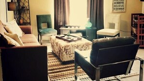 Un salón con sofá, sillón, reposapiés y una decoración de cabeza de ciervo. Se observa una pared con citas motivacionales y una ventana con cortinas.