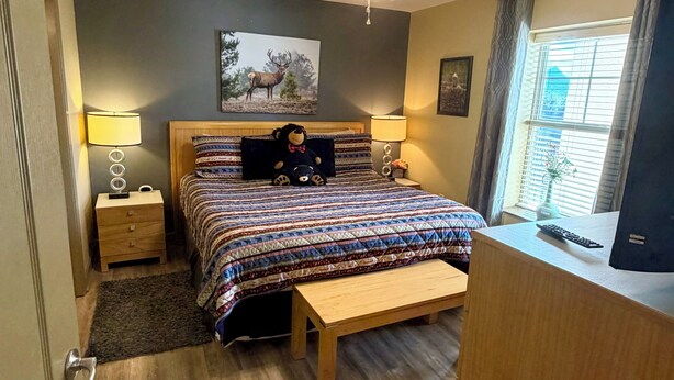 Ein Schlafzimmer mit einem Bett, Deckenventilator, Nachttisch mit Lampe, Fenster mit Vorhängen und Schreibtisch mit Tastatur.