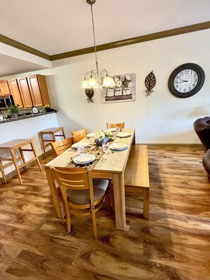Ein Essbereich mit einem Holztisch, gedeckt für vier Personen, umgeben von Stühlen. Im Hintergrund ist eine Küche mit Holzausstattung und einer Mikrowelle zu sehen. Über dem Tisch befinden sich eine Wanduhr und dekorative Gegenstände.