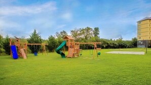 Ein Spielplatz mit Rutsche und Schaukeln, umgeben von einer grünen Wiese und Bäumen.