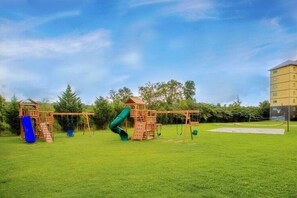 Ein Spielplatz mit Rutsche und Schaukeln, umgeben von einer grünen Wiese und Bäumen.