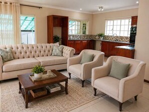 A living room with a sofa, two armchairs, a coffee table, and a kitchen in the background. - 2 Bed Garden Cottage |Pool| Garden (Sandton)