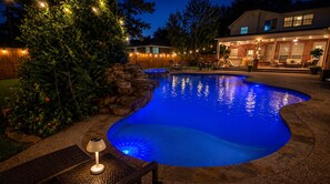 A swimming pool with a stone edge, surrounded by a wooden deck and a patio area with outdoor furniture.