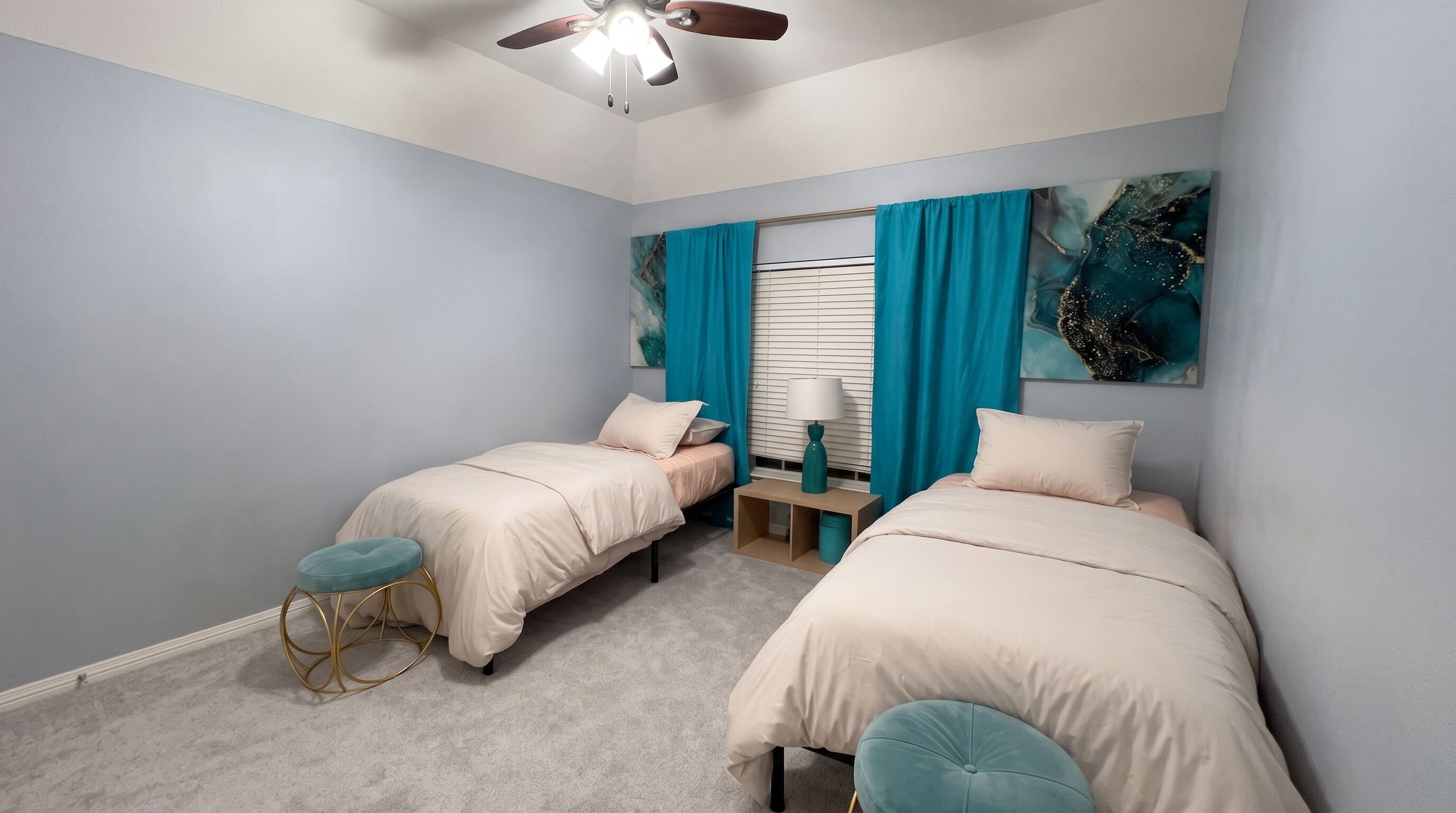 Une chambre avec deux lits, un ventilateur de plafond, une fenêtre avec des rideaux et un tableau au mur.