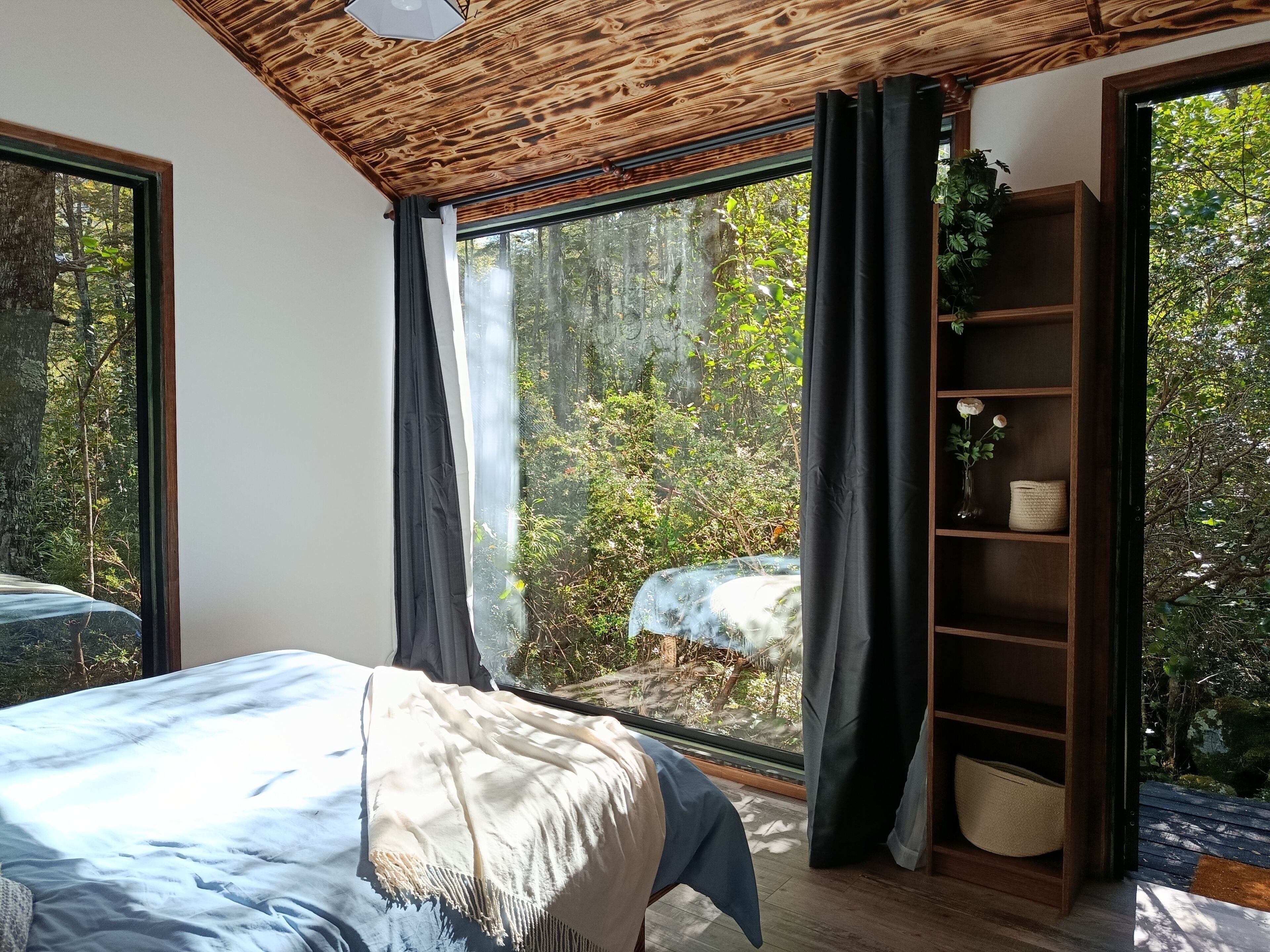Um quarto com uma janela grande, uma cama, uma estante de madeira e vista para uma floresta.