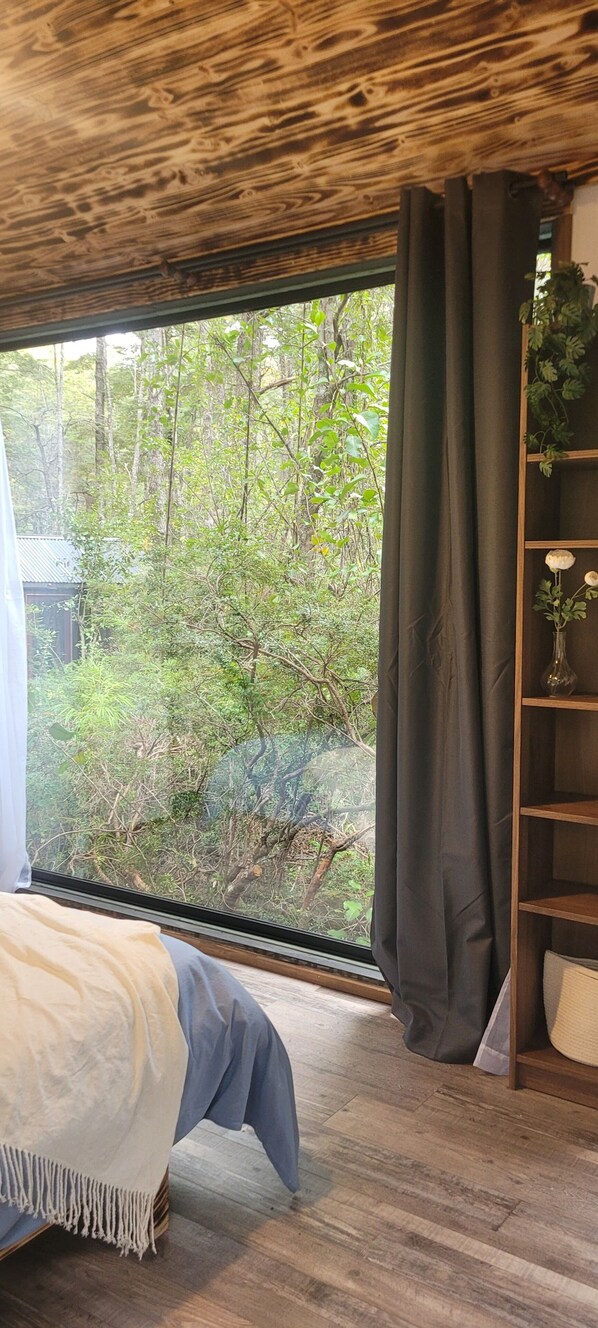 Een slaapkamer met een groot raam, houten plafond en een bed met een deken in wit en grijs.