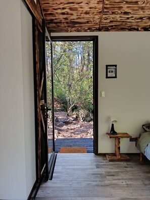 Ein Zimmer mit Holzbalkendecke, einem Bett mit grüner Bettdecke, einem Nachttisch mit Lampe und Blick auf eine natürliche Landschaft durch eine offene Tür.