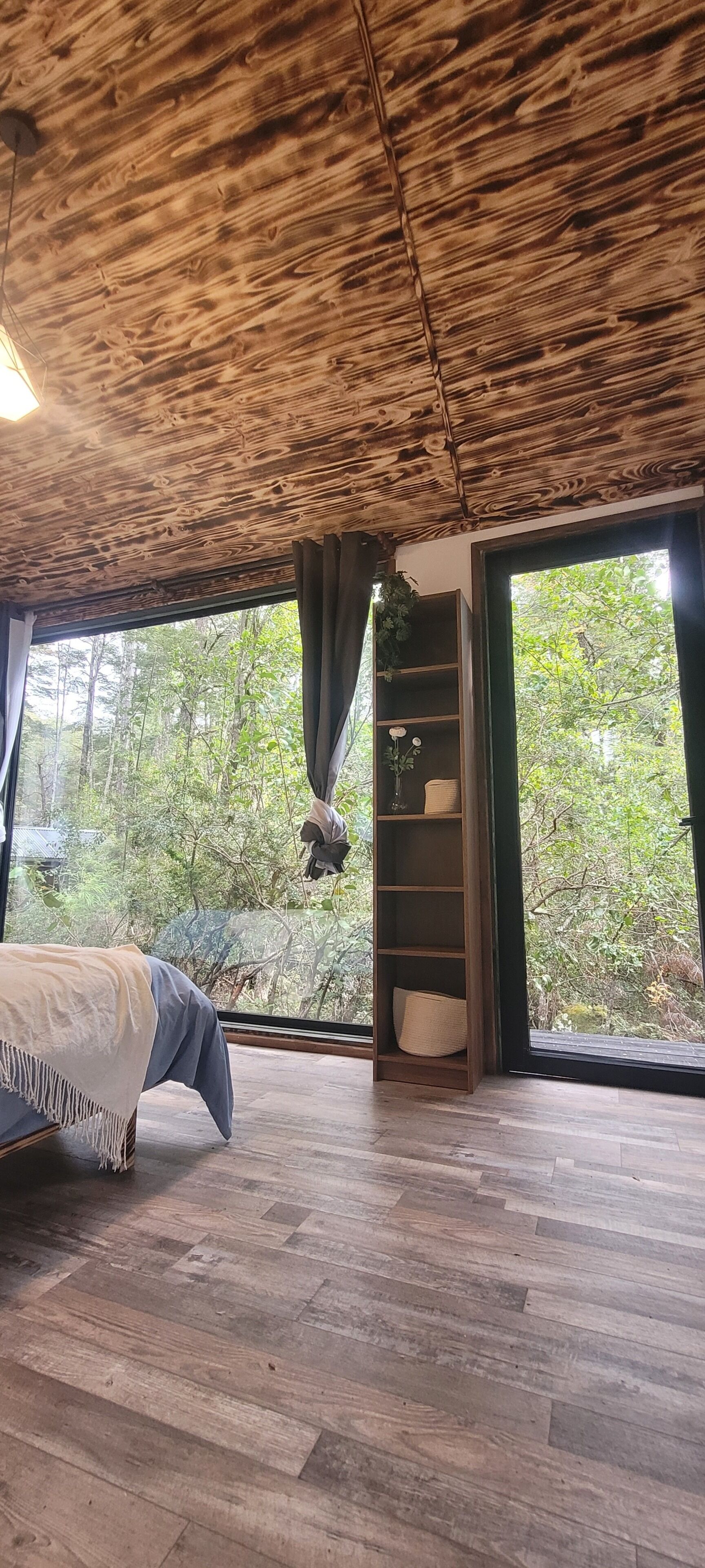 A room with a wooden ceiling, a bed, a window with curtains, and a shelf with a plant.