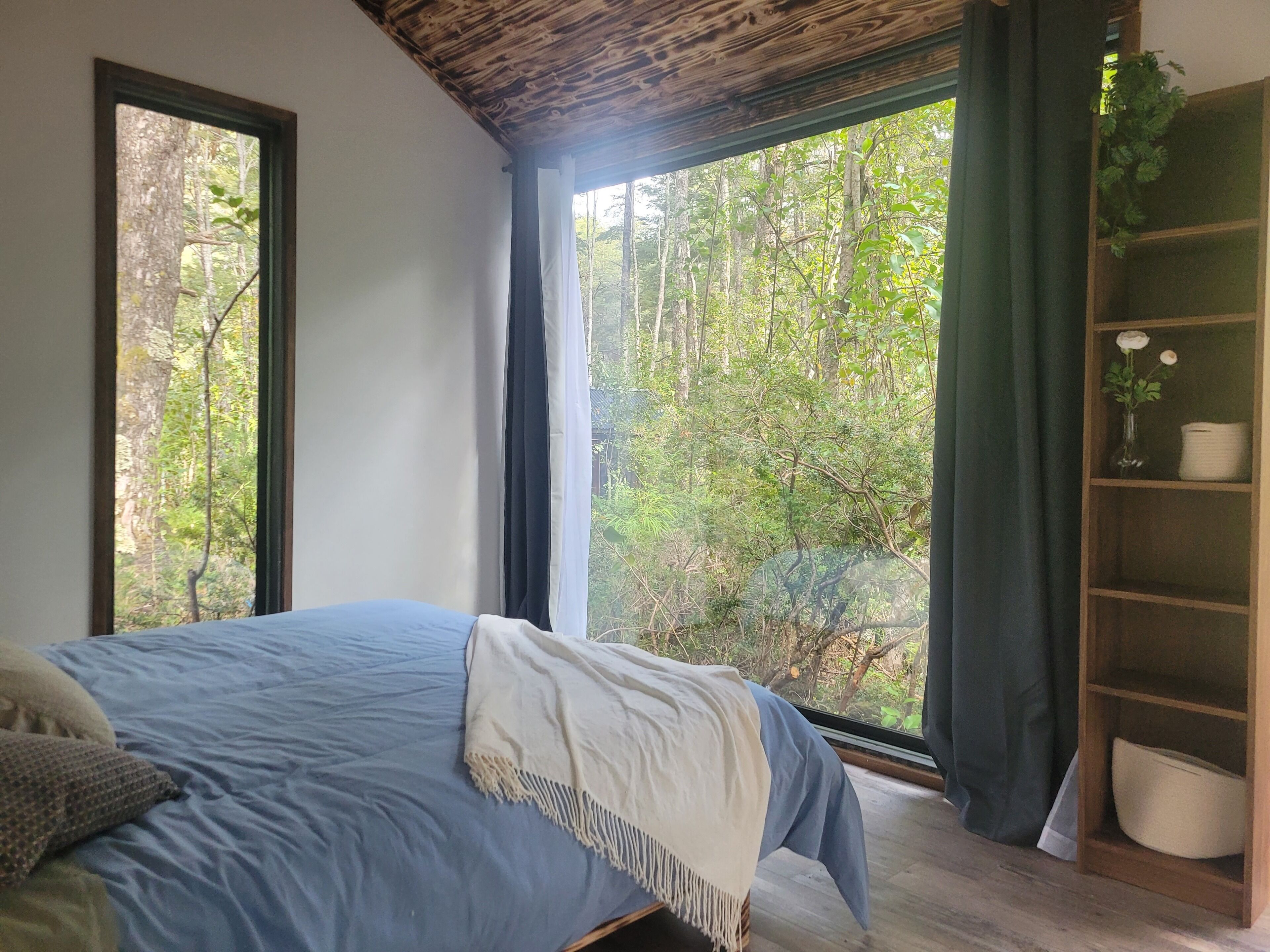 Ein Schlafzimmer mit großem Fenster, einem Bett mit blauer Bettwäsche und einem hölzernen Bücherregal.