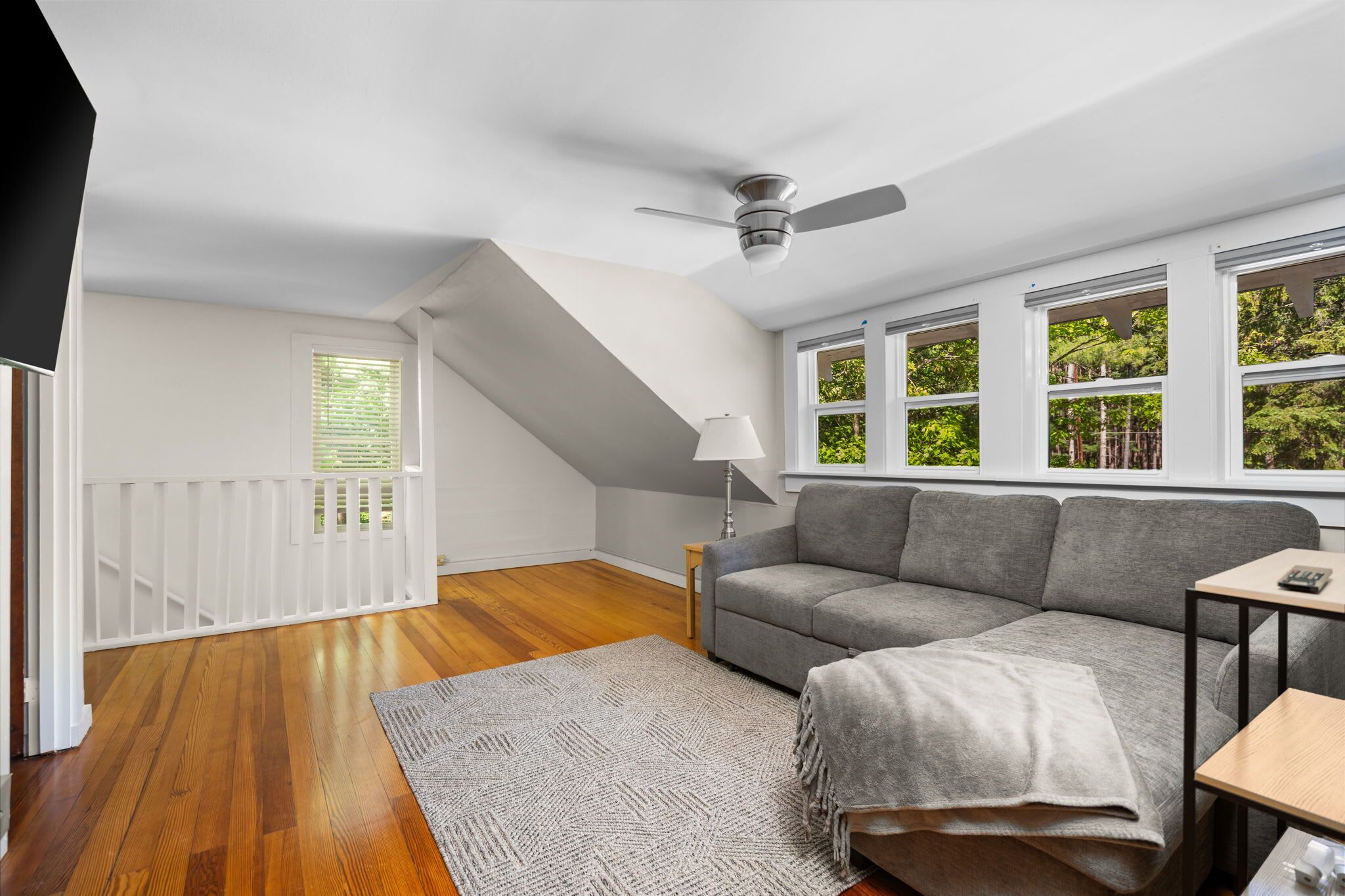 Ein modernes Wohnzimmer mit grauem Sofa, Deckenventilator und großen Fenstern mit Blick auf Grünflächen.