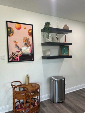 A room with a framed artwork of a leopard holding a martini glass, a small round table with a wicker chair, a trash can, and a set of black shelves with decorative items. - Cozy 2 bedroom home close to downtown Frisco! Enjoy all that Frisco has to offer (Frisco)