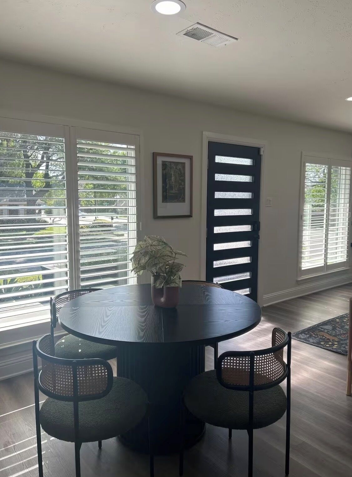 A dining area with a round table, four chairs, a potted plant, and a framed picture on the wall.