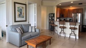 Una sala de estar con sofá, mesa de centro y una zona de cocina con refrigerador, taburetes de bar y microondas.