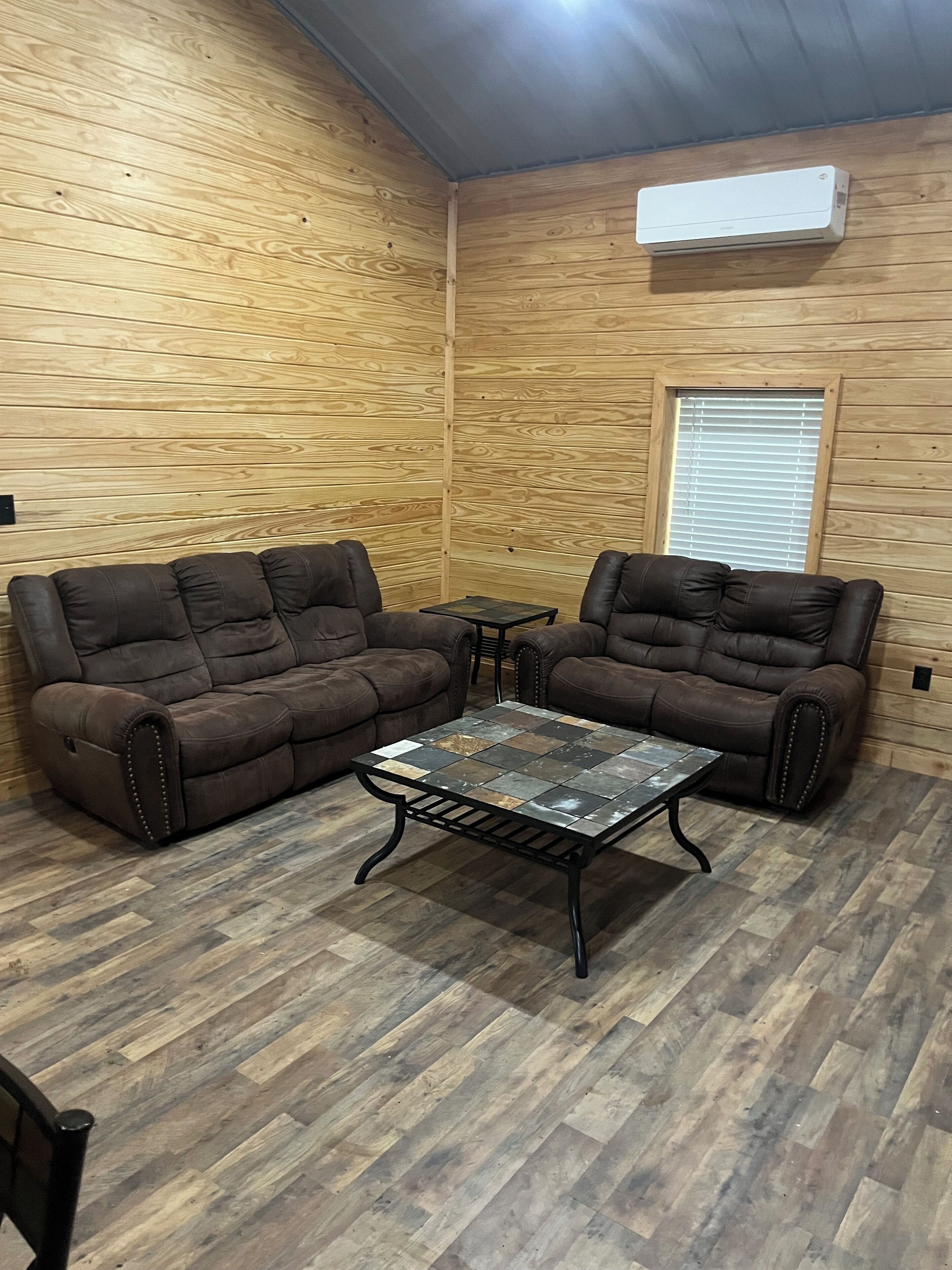 Una habitación con paredes de madera, dos sofás marrones, una mesita y un equipo de aire acondicionado.