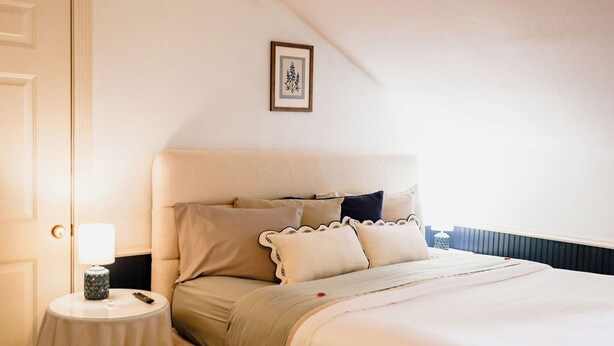 A bedroom with a bed, pillows, a bedside table with a lamp, and a framed picture on the wall. - The Woodrow House (Lubbock)