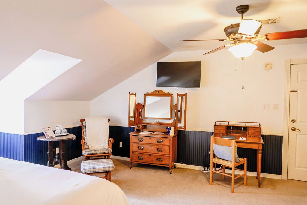 A bedroom with a bed, a desk, a chair, a dresser, a mirror, a ceiling fan, and a television on the wall.