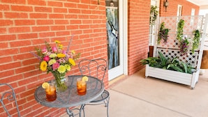 Un patio con una mesa redonda de metal, dos sillas, un jarrón con flores y una maceta con plantas.