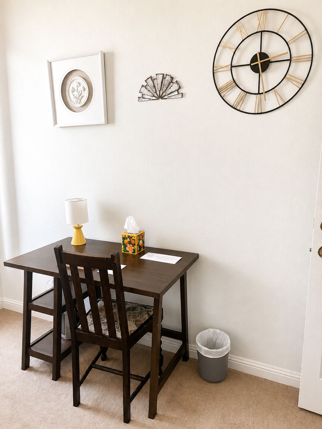 A dining area with a table, chairs, a wall clock, a framed picture, and a trash can.