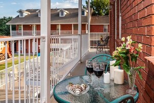 Una mesa de vidrio con una bandeja de fresas bañadas en chocolate, dos copas de vino y un florero con flores en un balcón con vista a una zona residencial.