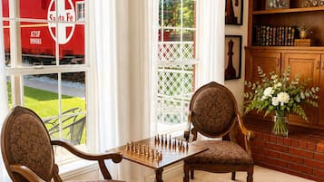 A room with a chessboard set on a table, two upholstered chairs, a bookshelf with books and decorative items, and a view of a red train outside.