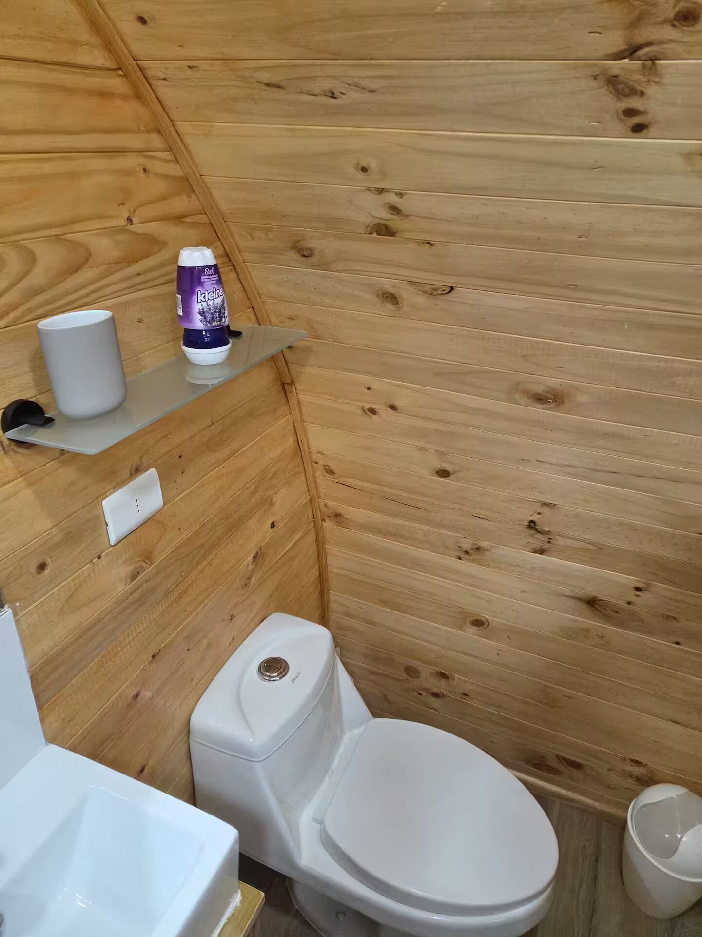 Une petite salle de bain en bois, avec des murs compacts, comprenant des toilettes, un lavabo blanc et une étagère sur laquelle sont posés un flacon et une tasse.