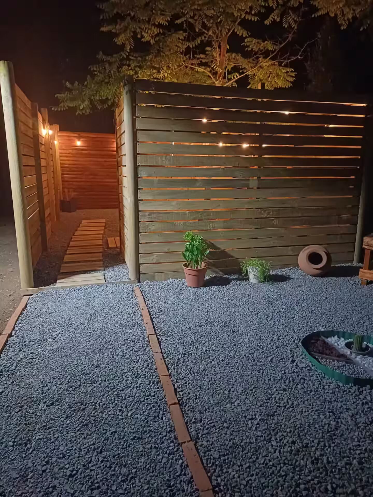 Una cerca de madera con tablones horizontales, un camino de grava y plantas en macetas.