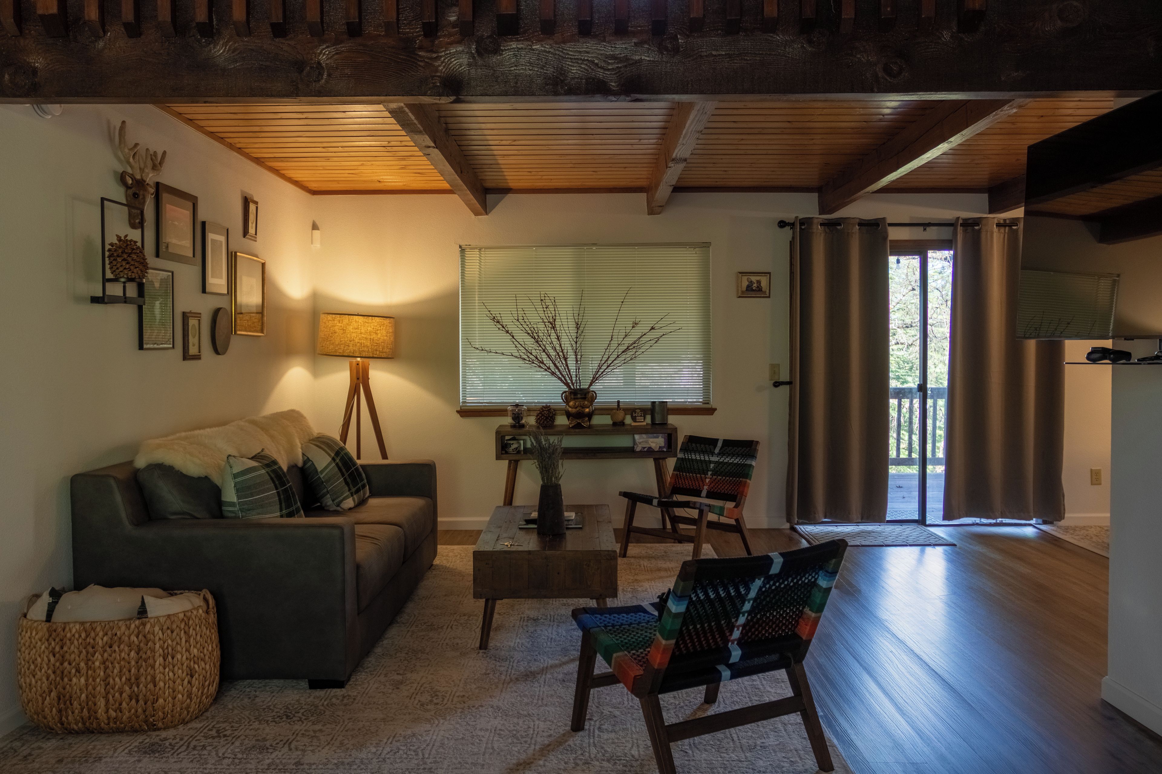 A cozy living room with a sofa, armchairs, a coffee table, and a lamp. There is a window with blinds and a door leading outside.