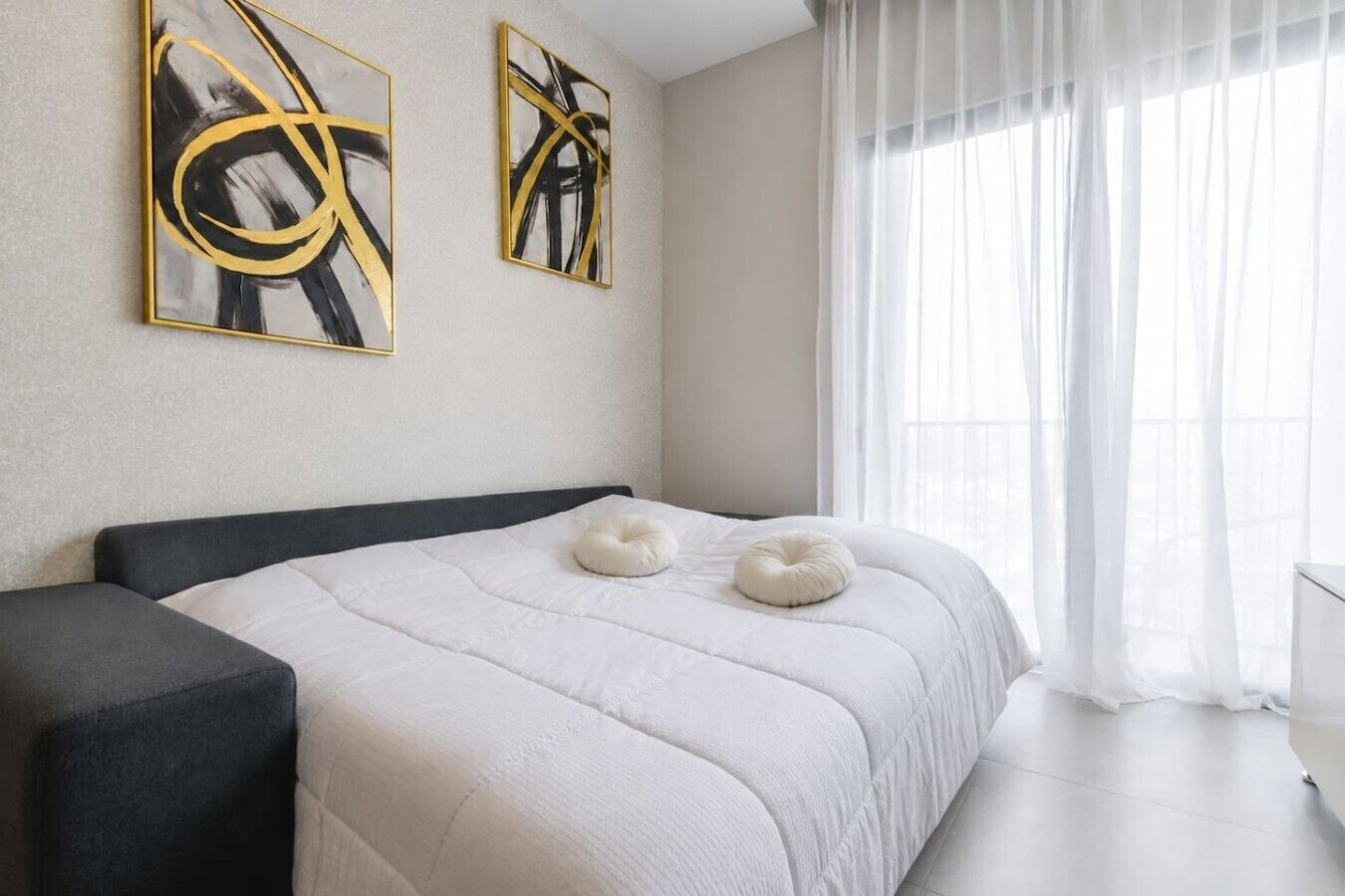 A modern bedroom with a bed, two abstract paintings, a window with sheer curtains, and a white pillow.