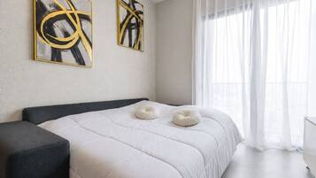 A modern bedroom with a bed, two abstract paintings, a window with sheer curtains, and a white pillow.