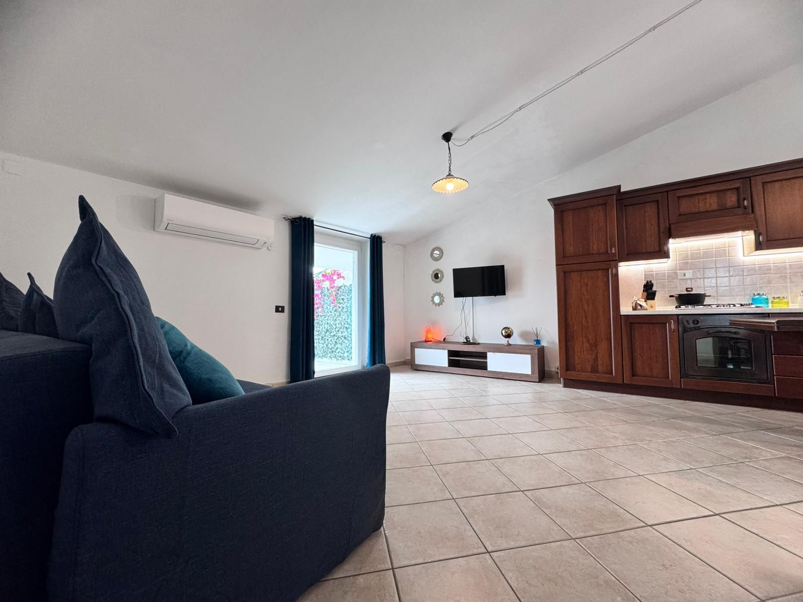 Un saló modern amb un sofà, una televisió plana i una cuina amb armaris de fusta i una placa de cocció.