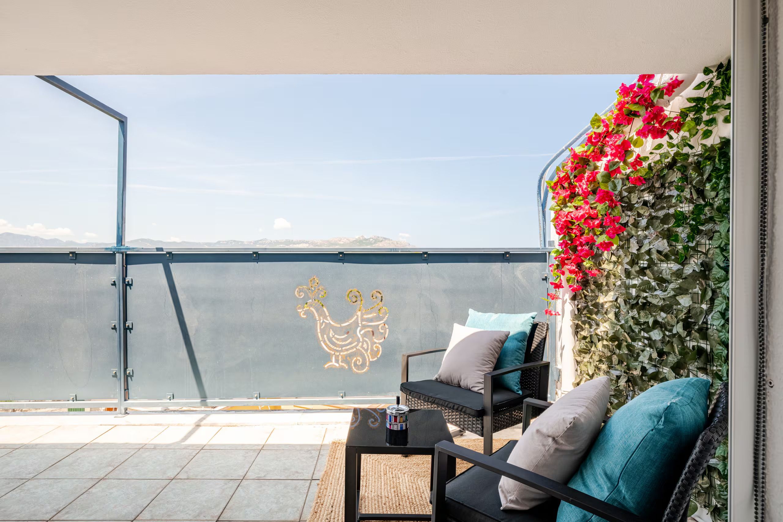 A balcony with a view of the sea, a decorative wall with flowers, and a seating area with cushions.