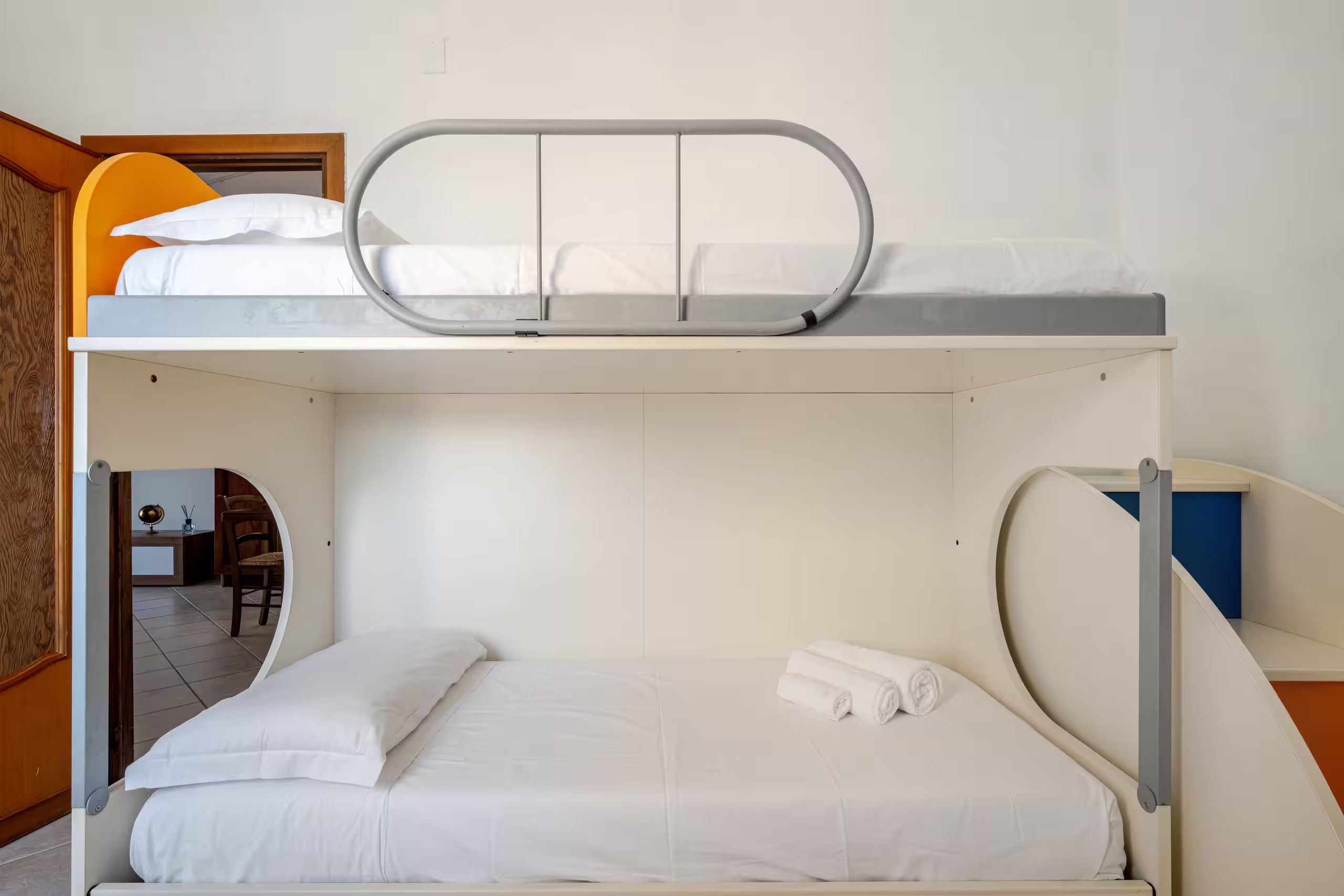 Una cama litera con colchón y almohadas, una mesita con una lámpara y una silla en la habitación contigua.