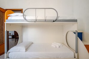 A bunk bed with a mattress and pillows, a small table with a lamp, and a chair in the adjacent room.